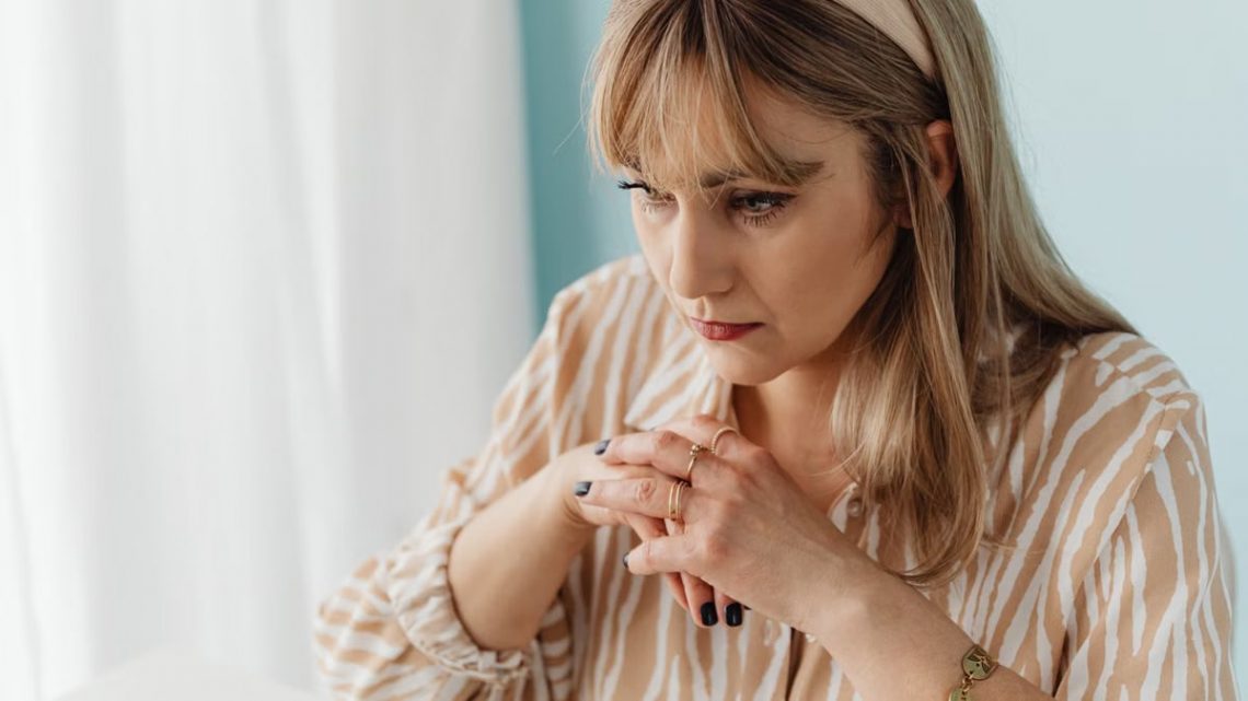 Pensive woman indoors, hands clasped, looking contemplative. Captured in natural light.